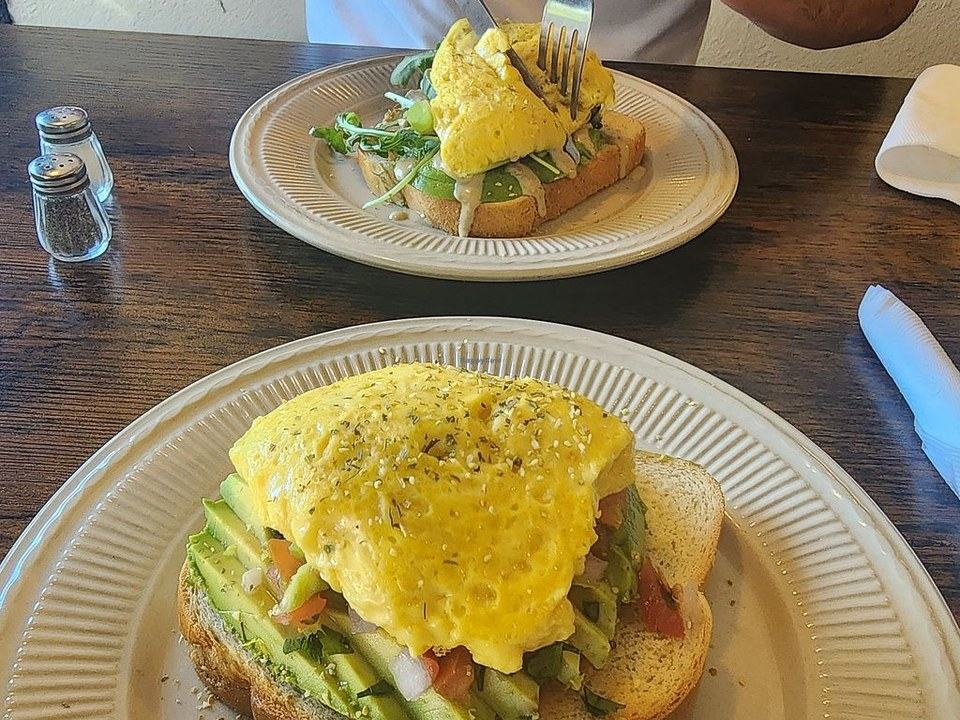 Two plates of omelette on toast. The near plate has avocado, tomato, and onion under the eggs.