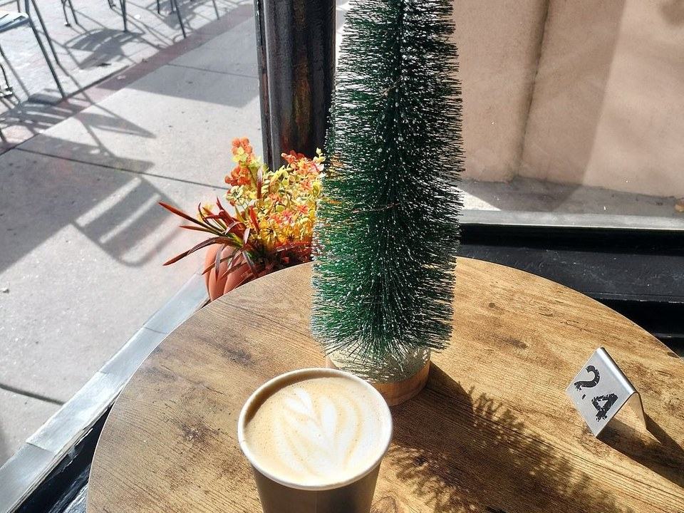 A latte with leaf art in a paper cup on a wood table by the front window, next to a bottle-brush tree and table number 24.